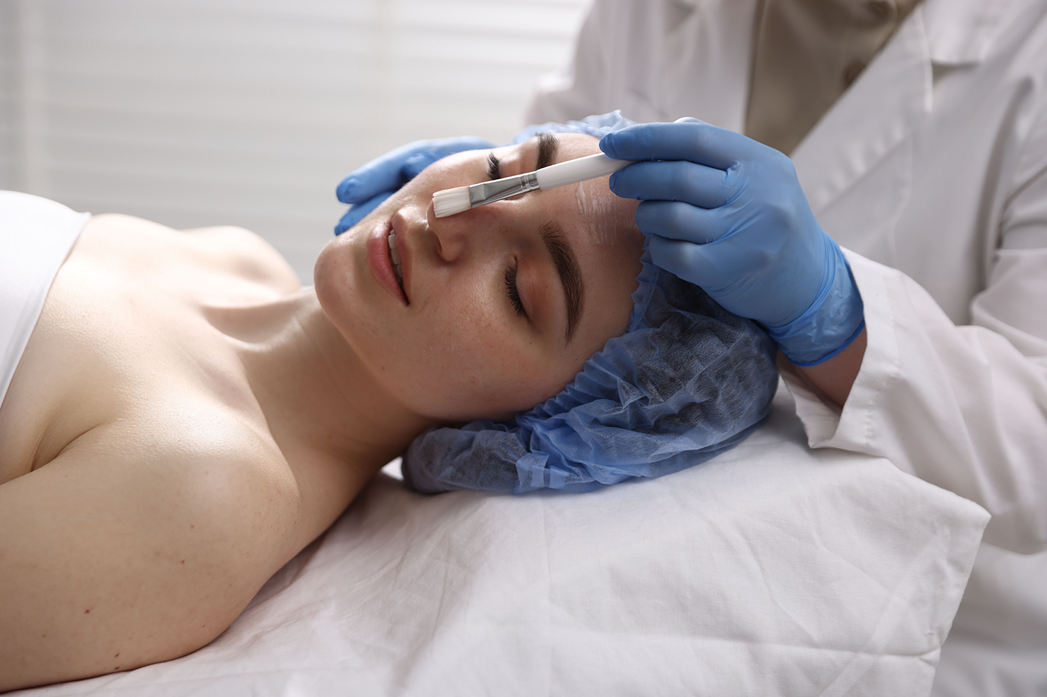 Chemical Peel Facial Esthetics in Orange Park Woman lying back with eyes closed and a blue cap on her hair while a provider in blue gloves brushes a chemical peel onto her face, shown for facial esthetics in Orange Park