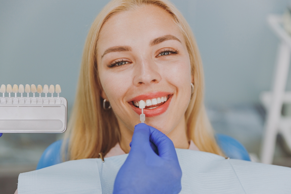 Smiling woman with long blonde hair reclined in a dental chair while a gloved hand holds a tooth shade sample beside her teeth to choose a veneer color, shown for porcelain veneers in Orange Park