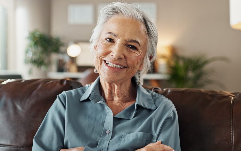 Smiling older woman with short gray hair and a gray-blue collared shirt relaxing on a brown leather sofa in a bright, comfortable room, representing bone grafting care in Orange Park