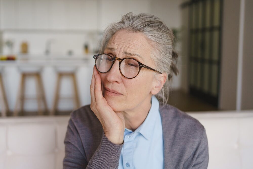 Root Canal Treatment for Tooth Pain in Orange Park Older woman with gray hair and tortoiseshell glasses resting her hand on her cheek with a pained look, showing tooth pain that may call for root canal treatment in Orange Park